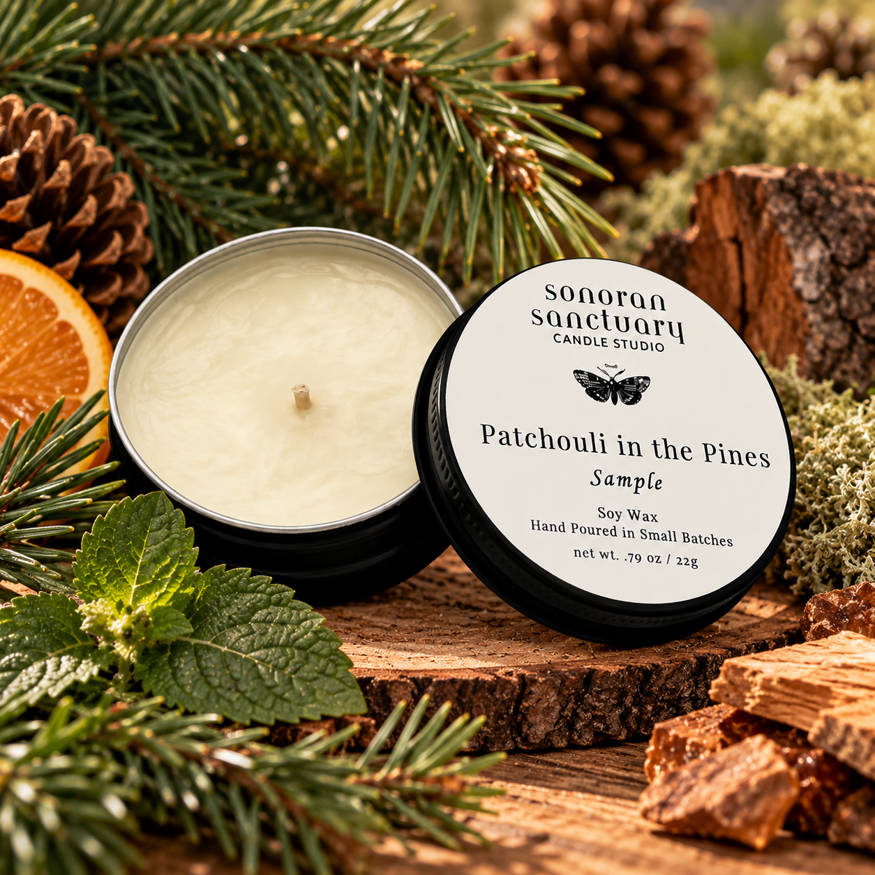 Sonoran Sanctuary Patchouli in the Pines soy candle sample tin with branded lid, surrounded by fresh pine branches, pinecones, patchouli leaves and natural wood